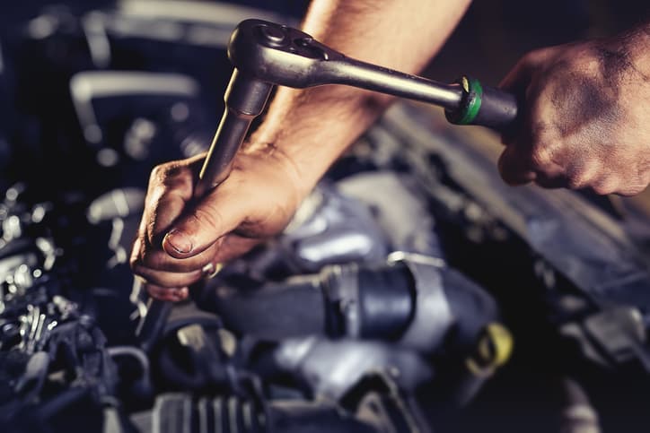 A mechanic uses a ratchet wrench while working on a vehicle engine. The image represents professional automotive repair, engine diagnostics, routine maintenance, and mechanical repair services performed by an experienced technician.